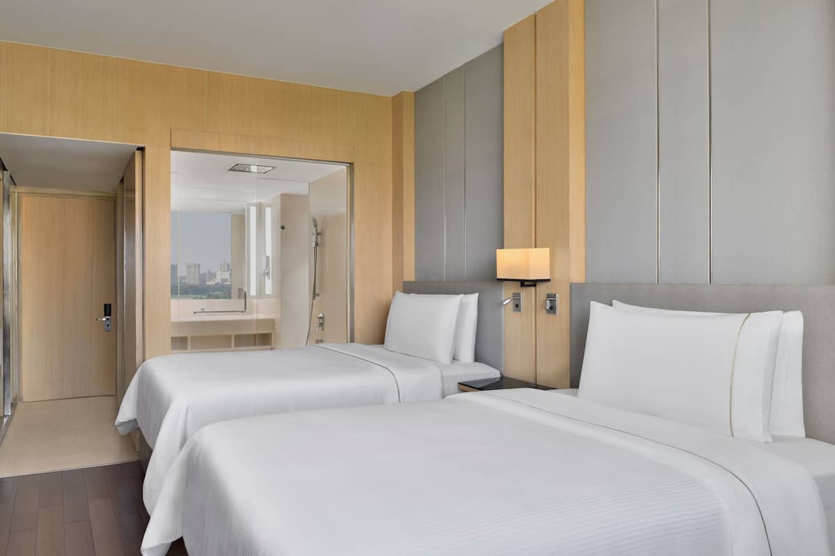 A hotel room with two beds, a wooden panel wall, a grey headboard, a bedside table with a lamp, and a view of the city through an open door.