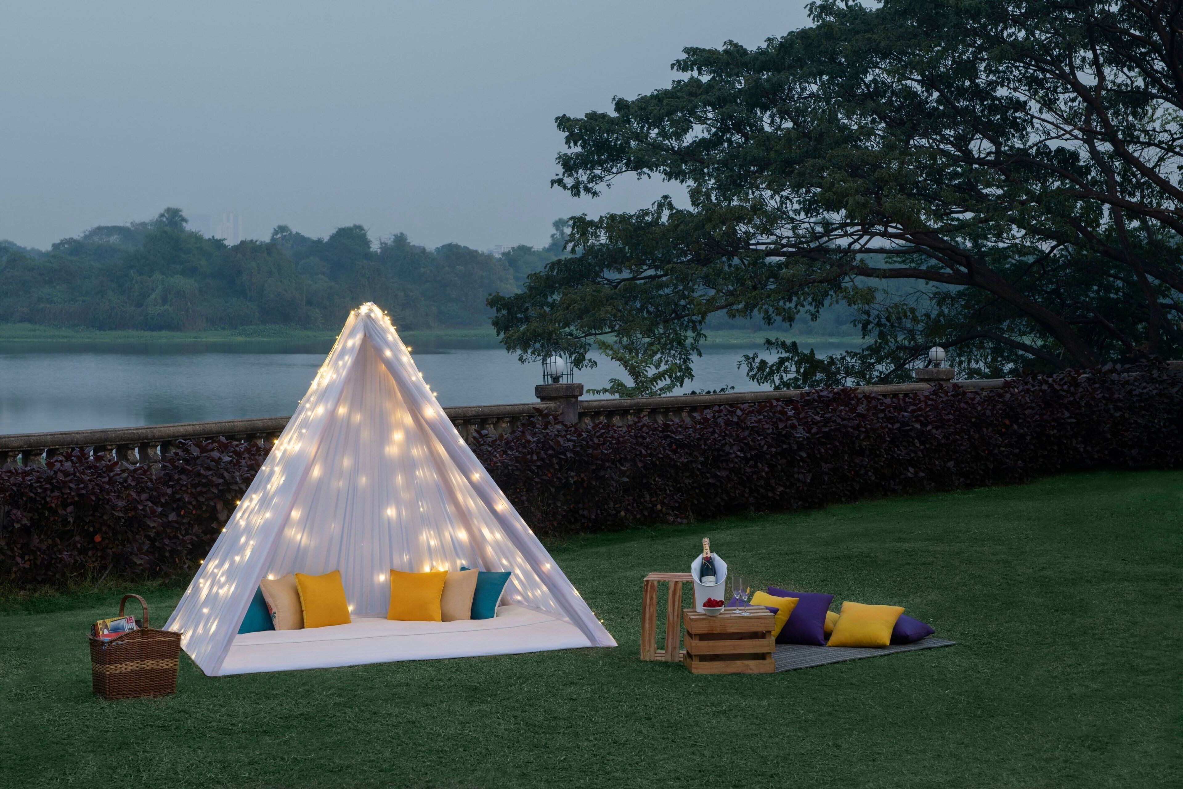 A tent with string lights, a picnic setup with a basket and crate, and a view of a lake and trees.