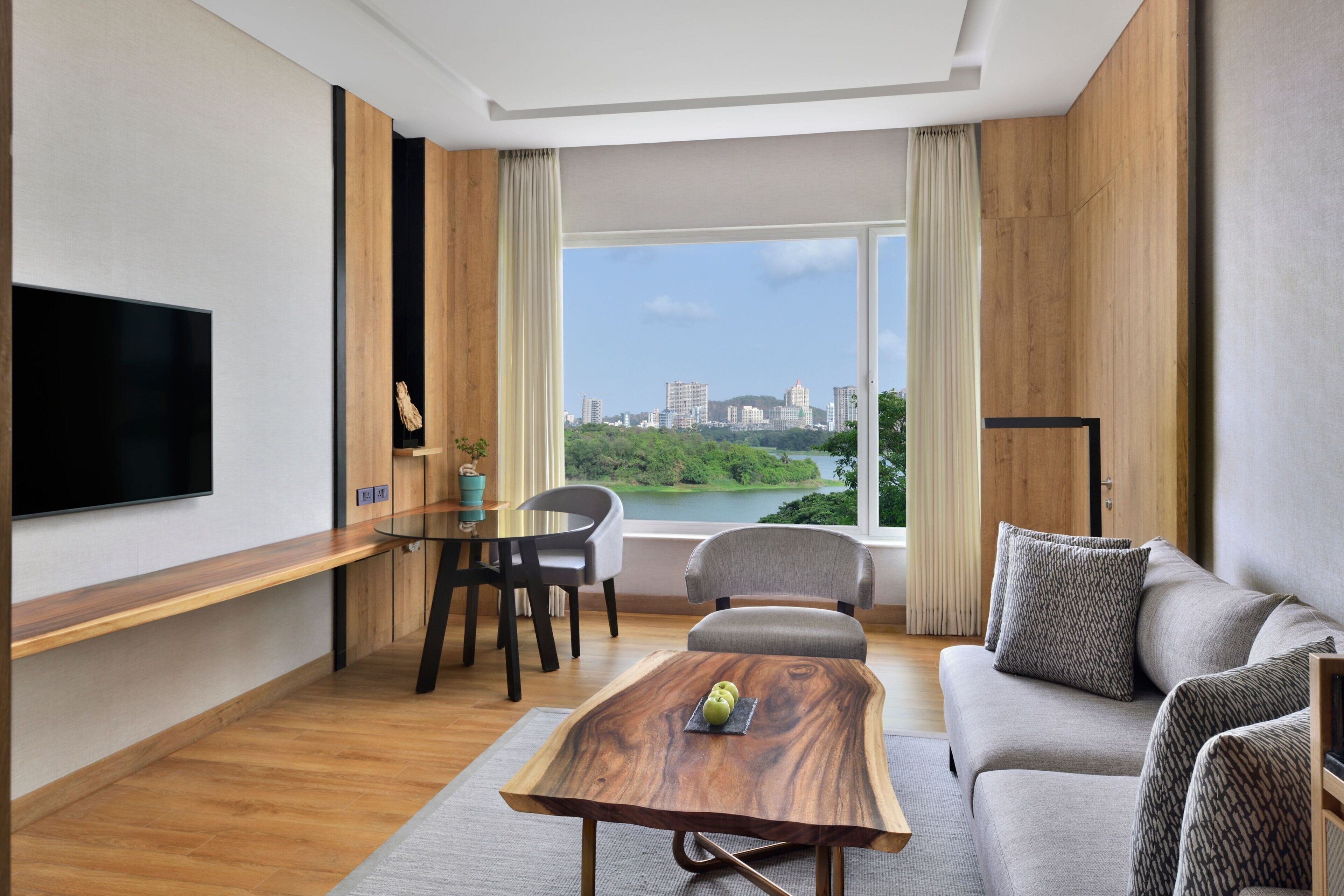 A modern living room with a flat-screen TV, a wooden coffee table, a sofa, and a view of a cityscape through large windows.