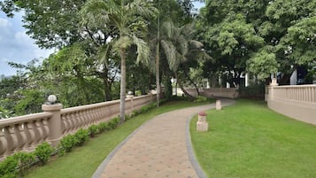 Un camino bordeado por una barandilla color beige y abundante vegetación, que conduce a un edificio con fachada blanca y techo curvo.
