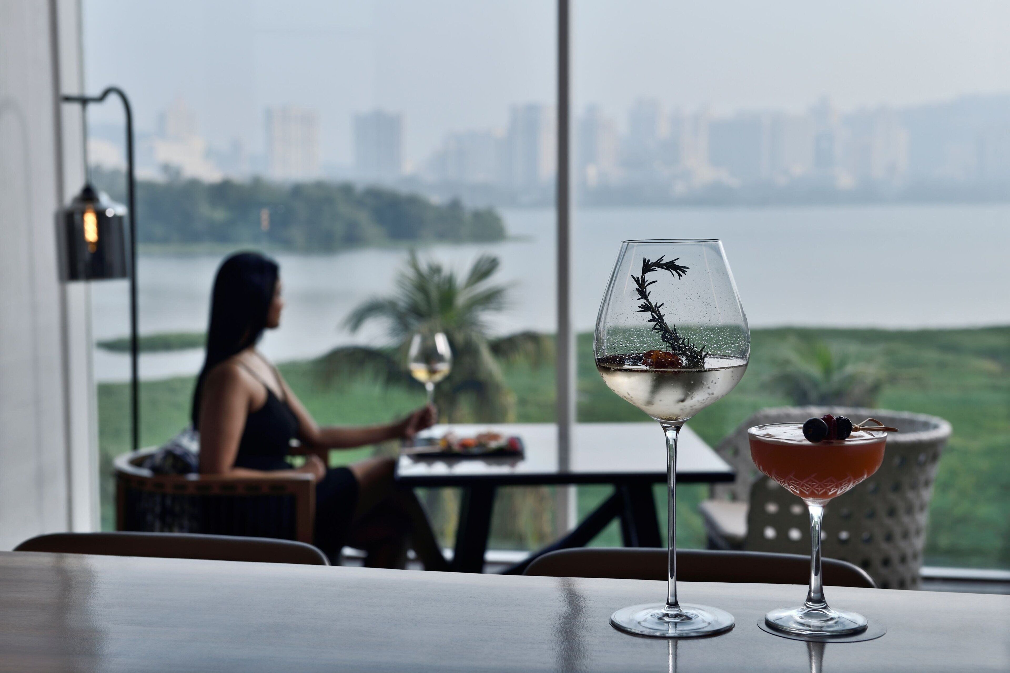 A table with two glasses of drink, a city view, and a person sitting at a table.
