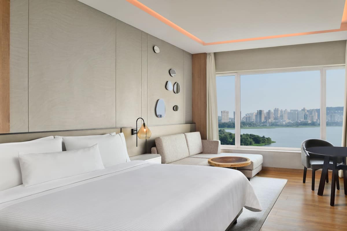 A modern hotel room with a large bed, a sofa, a dining area, and a view of a cityscape through large windows.