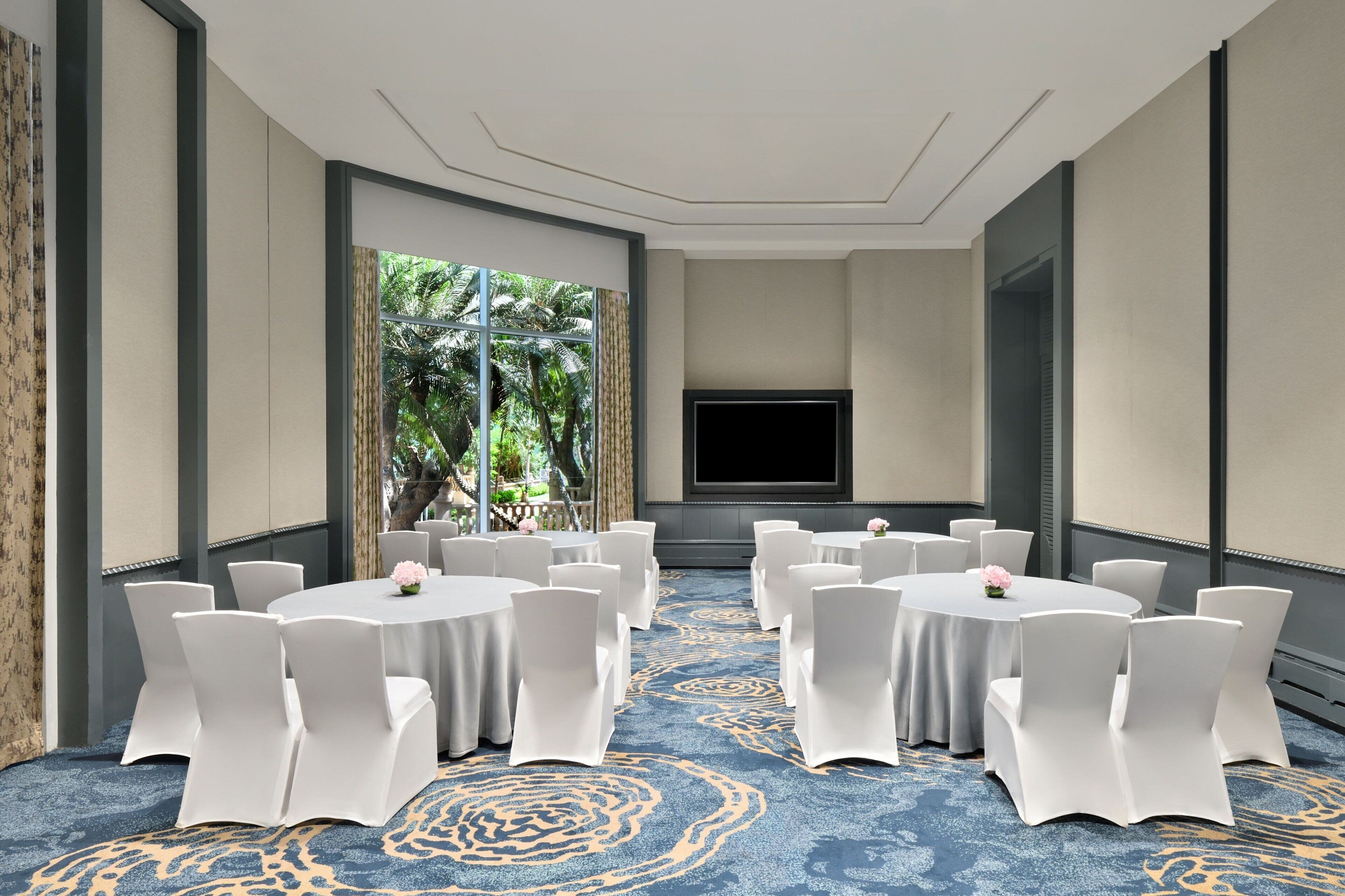 A conference room with round tables and white chairs, a large window with a view of greenery, and a flat-screen TV mounted on the wall.