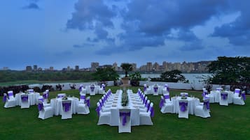 An outdoor wedding setup with tables and chairs arranged on a grassy area, overlooking a body of water and a city skyline.