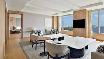 A modern hotel room with a large window, a sofa, a dining table, and a flat-screen TV.