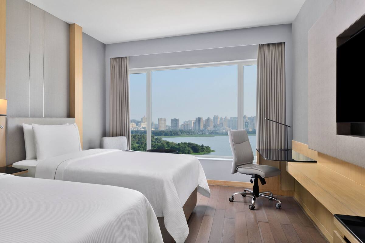 A hotel room with two beds, a desk, a chair, and a large window offering a city view.