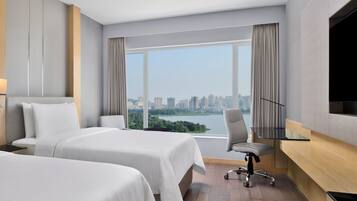 A hotel room with two beds, a desk, a chair, and a large window offering a city view.