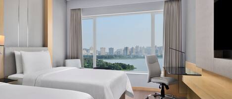 A hotel room with two beds, a desk, a chair, and a large window offering a city view.