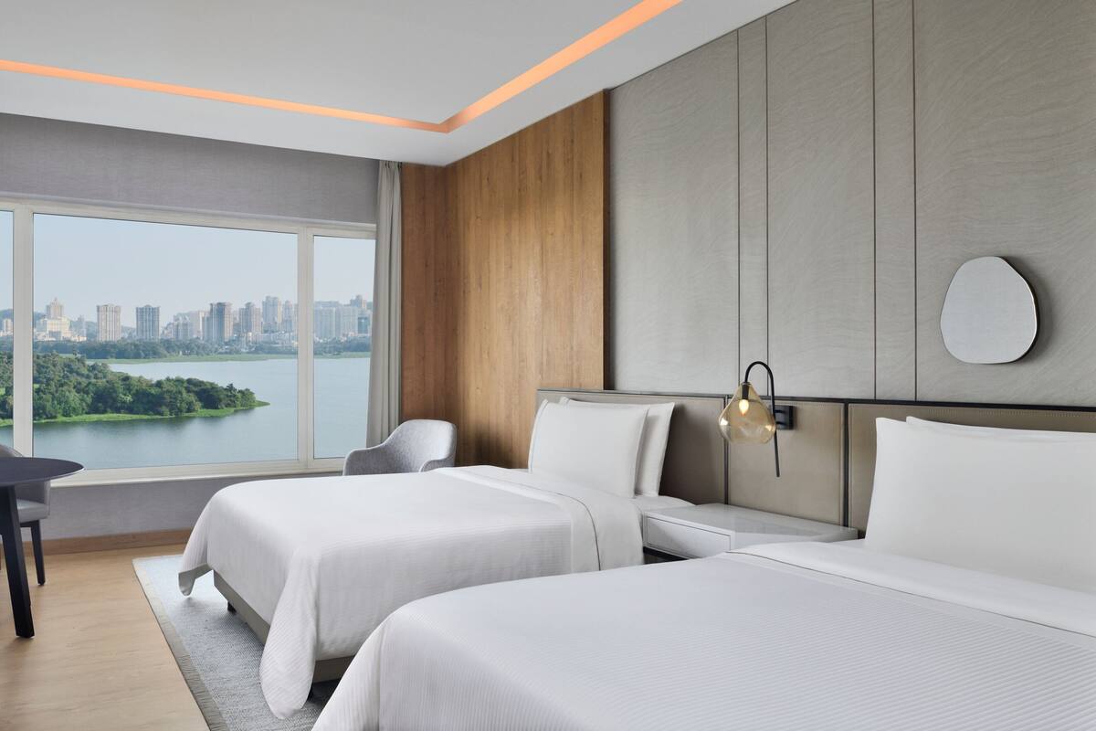 A modern hotel room with two beds, a view of a cityscape and a lake, a desk, and a chair.