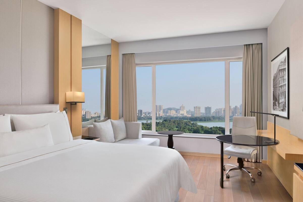 A hotel room with a large bed, a desk, a chair, and a view of a cityscape through large windows.