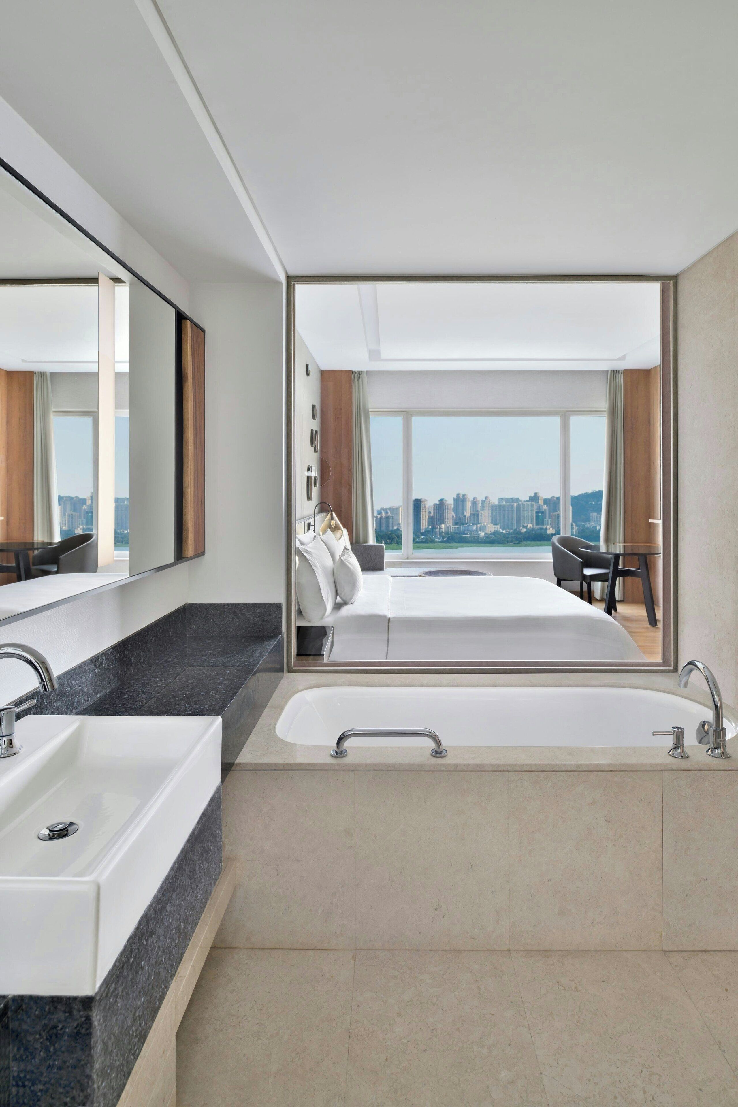 A modern hotel room with a large bed, a separate bathroom with a bathtub, a large mirror, and a city view.
