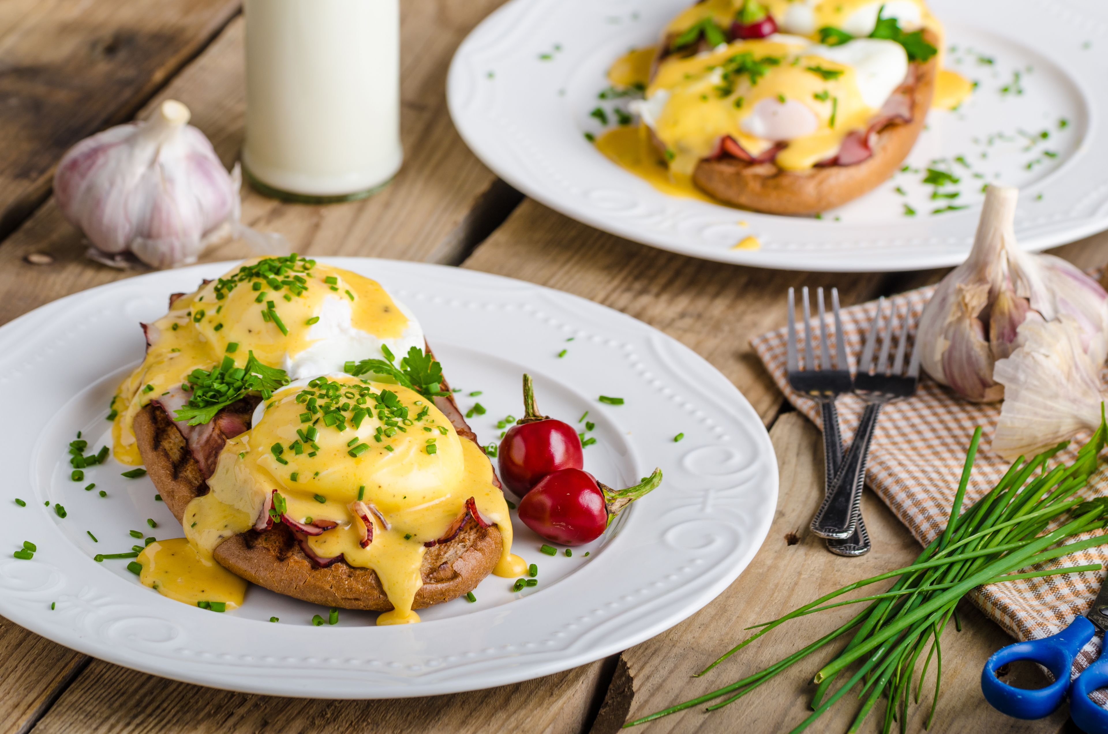 A breakfast setting with eggs Benedict on white plates, garnished with chives, and surrounded by garlic bulbs and chives.