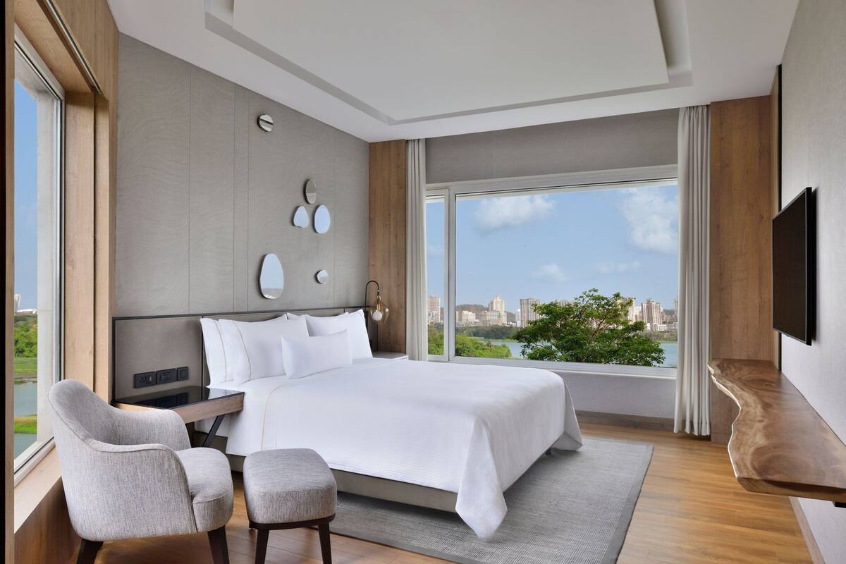 A modern hotel room with a large bed, a seating area with a chair and ottoman, a flat-screen TV mounted on the wall, and a large window offering a city view.
