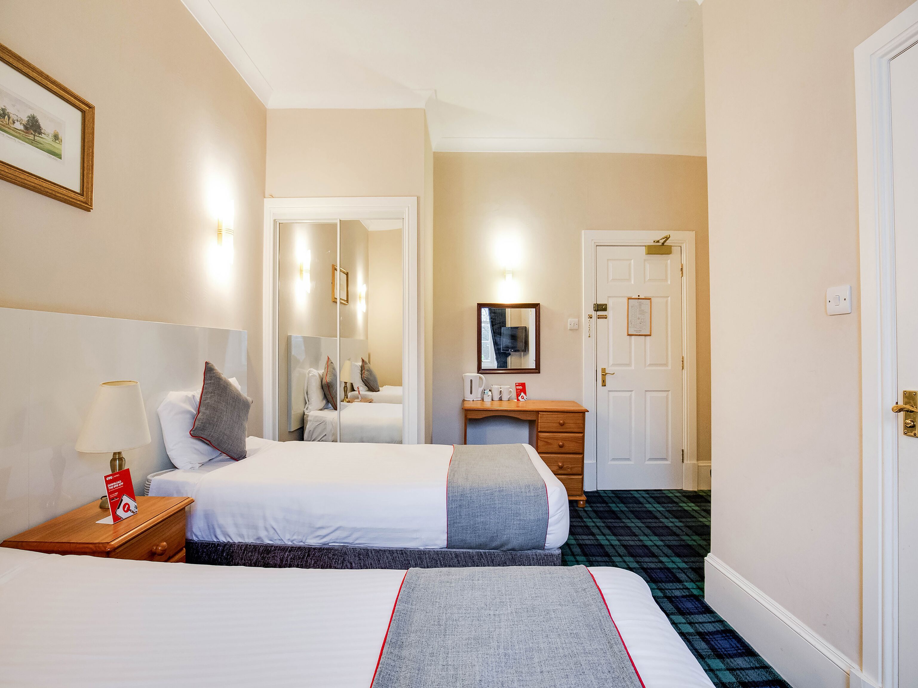 Habitación de hotel con dos camas, una mesita de noche con lámpara, cómoda, espejo y una puerta con cerradura.