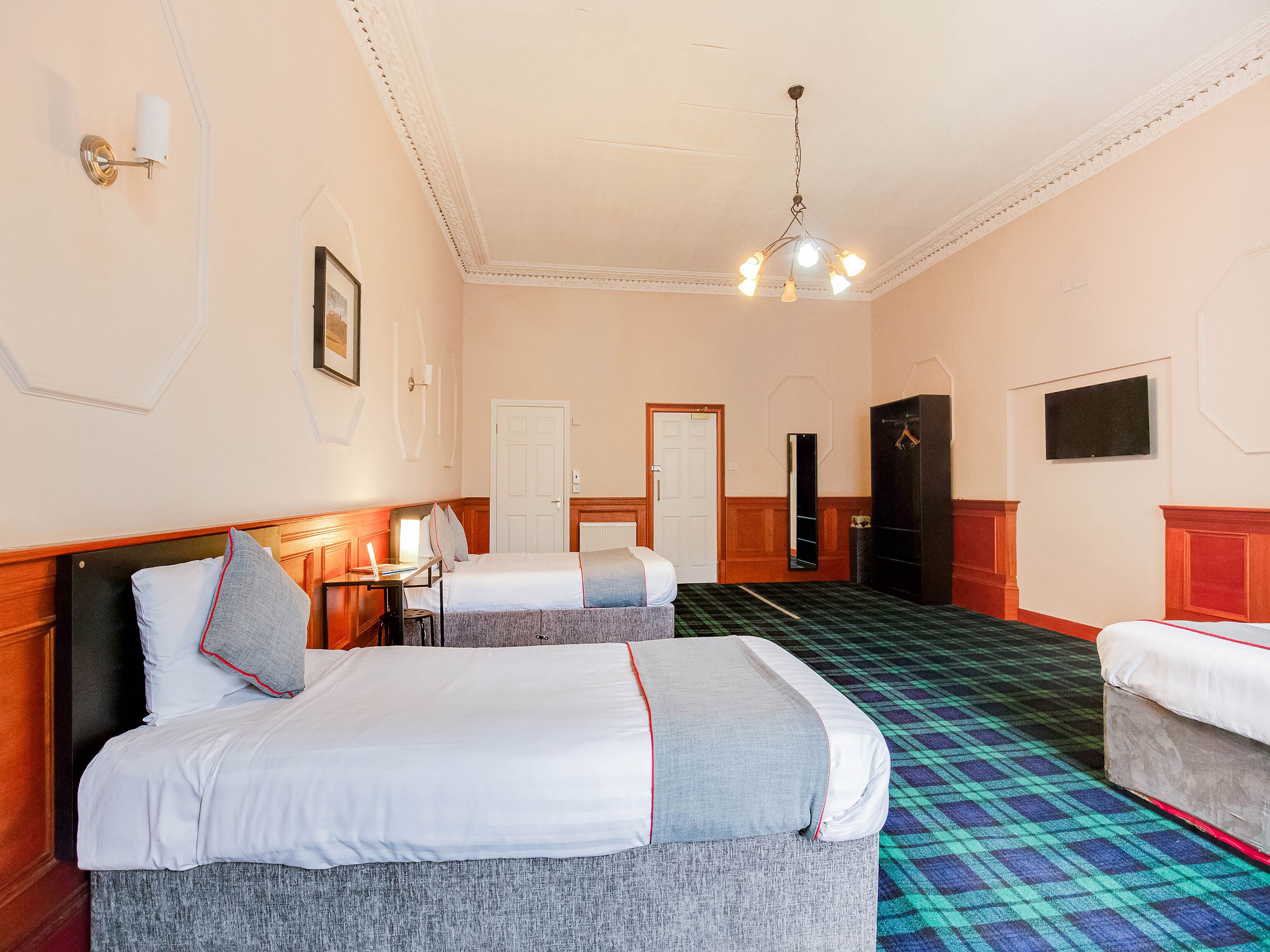 Habitación de hotel con dos camas, un escritorio, una silla, un armario, un espejo, un televisor y una alfombra estampada.
