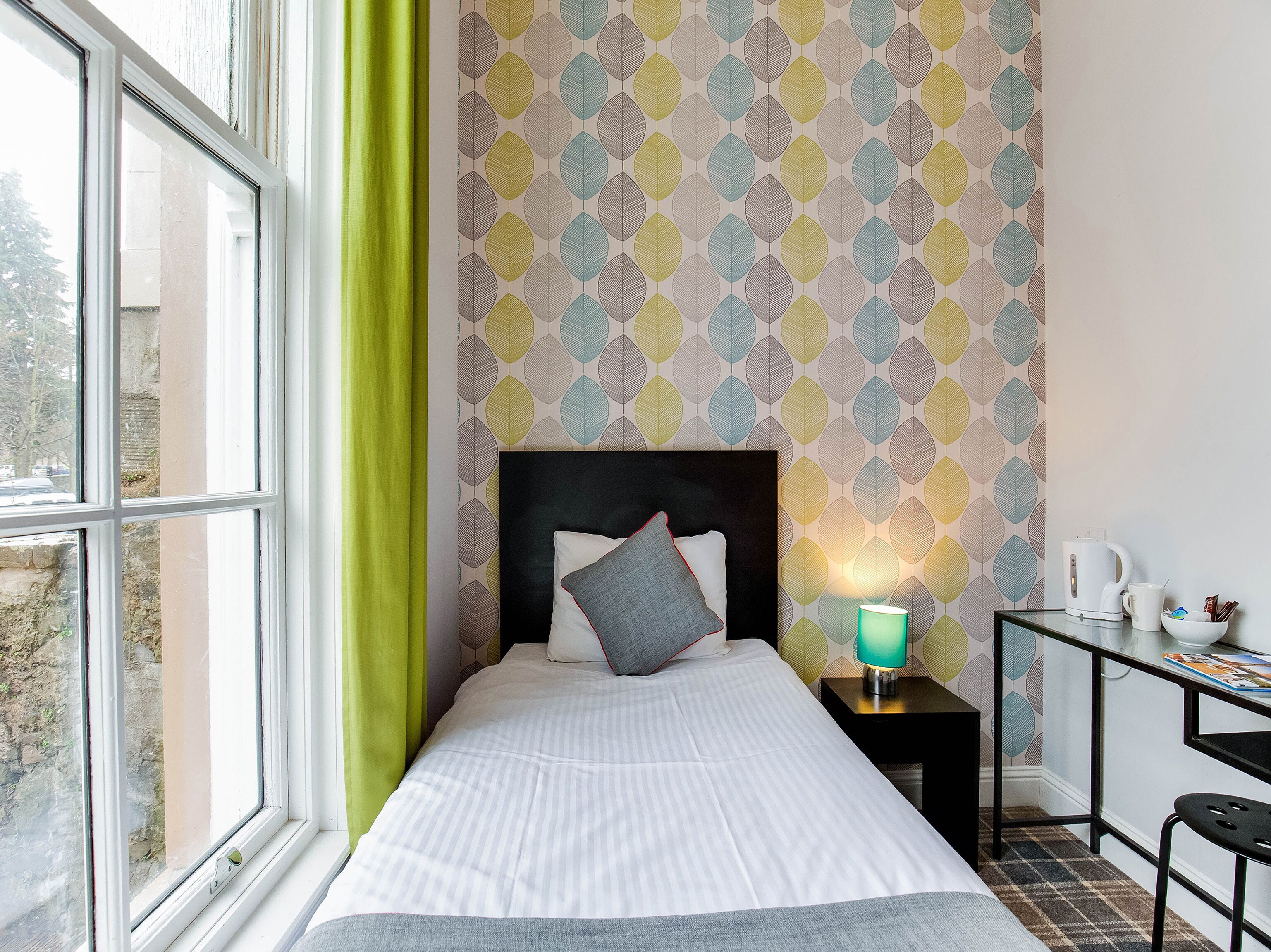 A neatly made bed with pillows, a bedside table with a lamp, a small desk with a chair, and a window with a green curtain.
