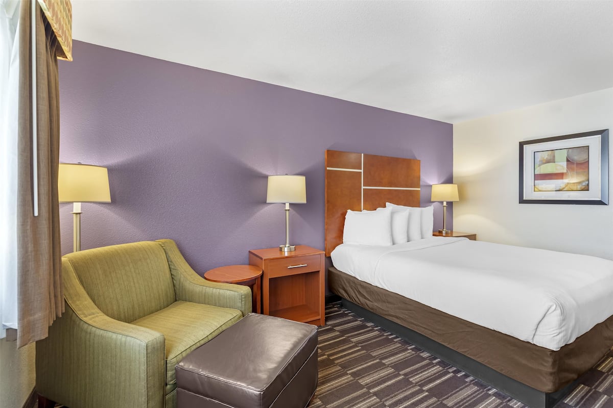 A hotel room with a bed, a green armchair, a small table, a nightstand with a lamp, and a wall-mounted painting.