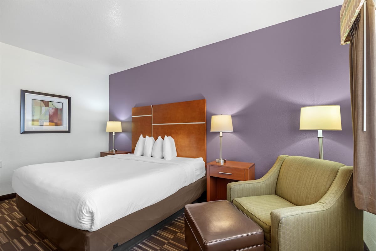 A hotel room with a large bed, two bedside tables with lamps, a green armchair, and a brown ottoman.