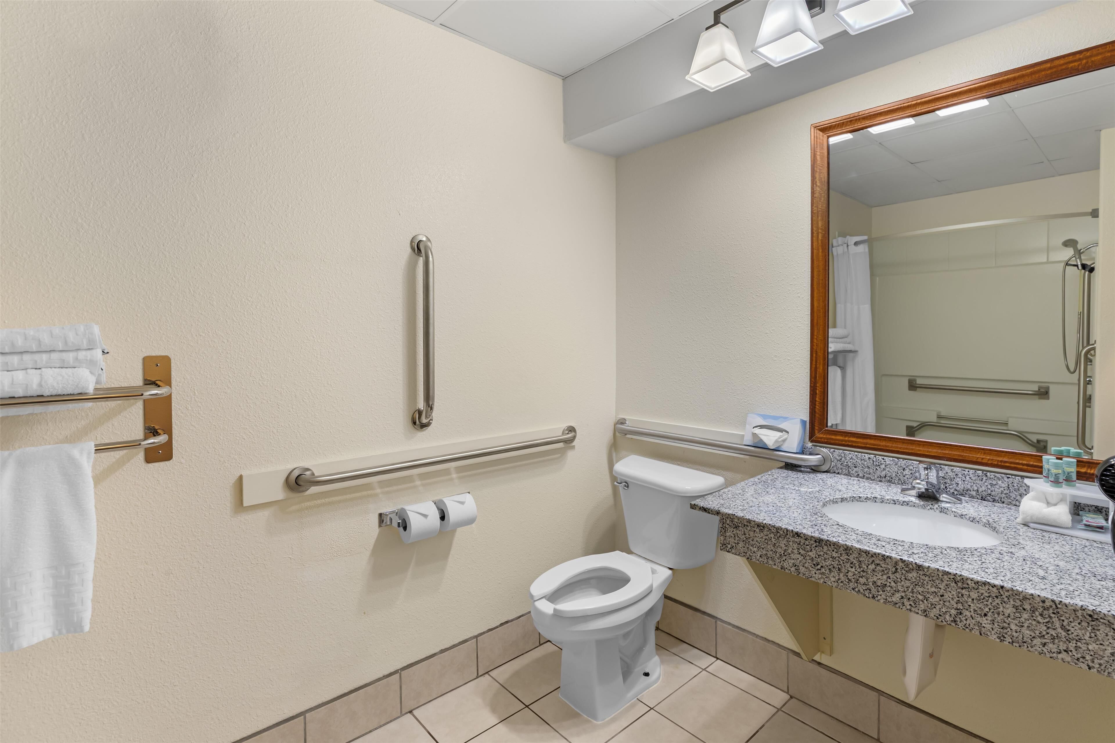 Une salle de bain avec des toilettes blanches, un lavabo, un miroir et des barres d’appui.