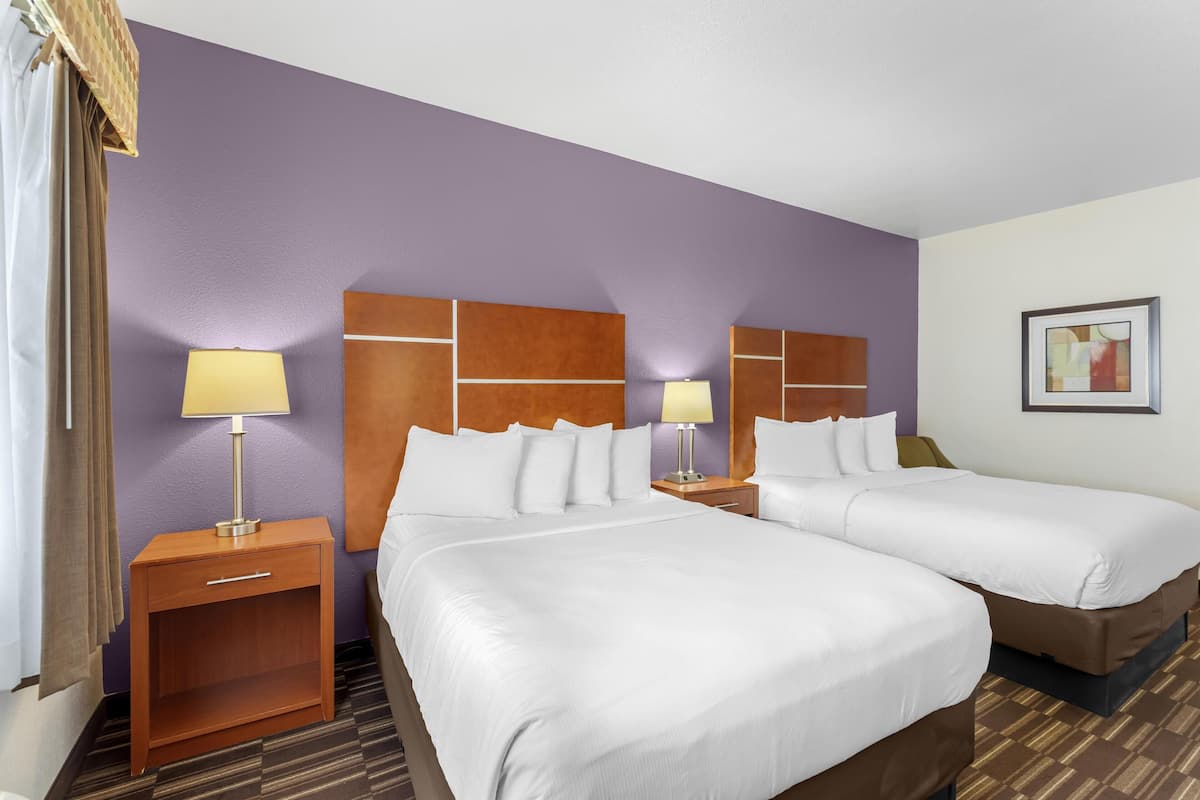 A hotel room with two beds, a wooden nightstand with a lamp, a small table, and a framed picture on the wall.