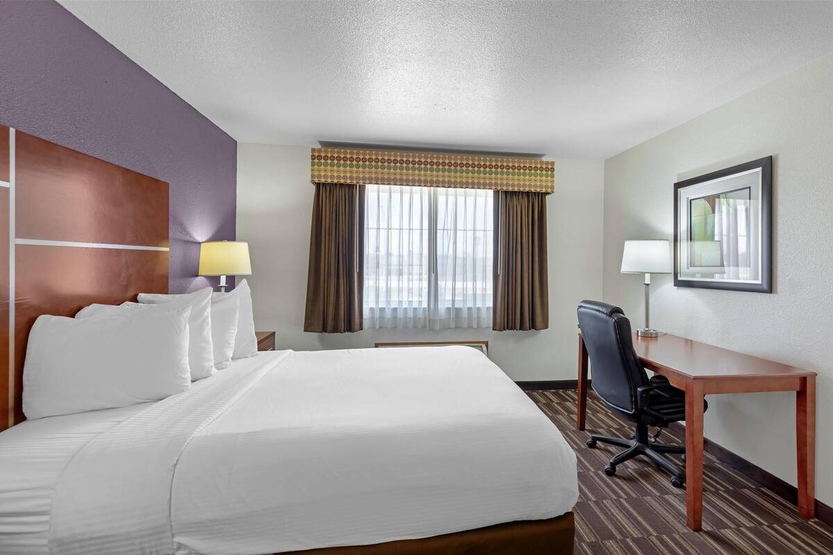 A hotel room with a large bed, a desk, a chair, and a window with curtains.