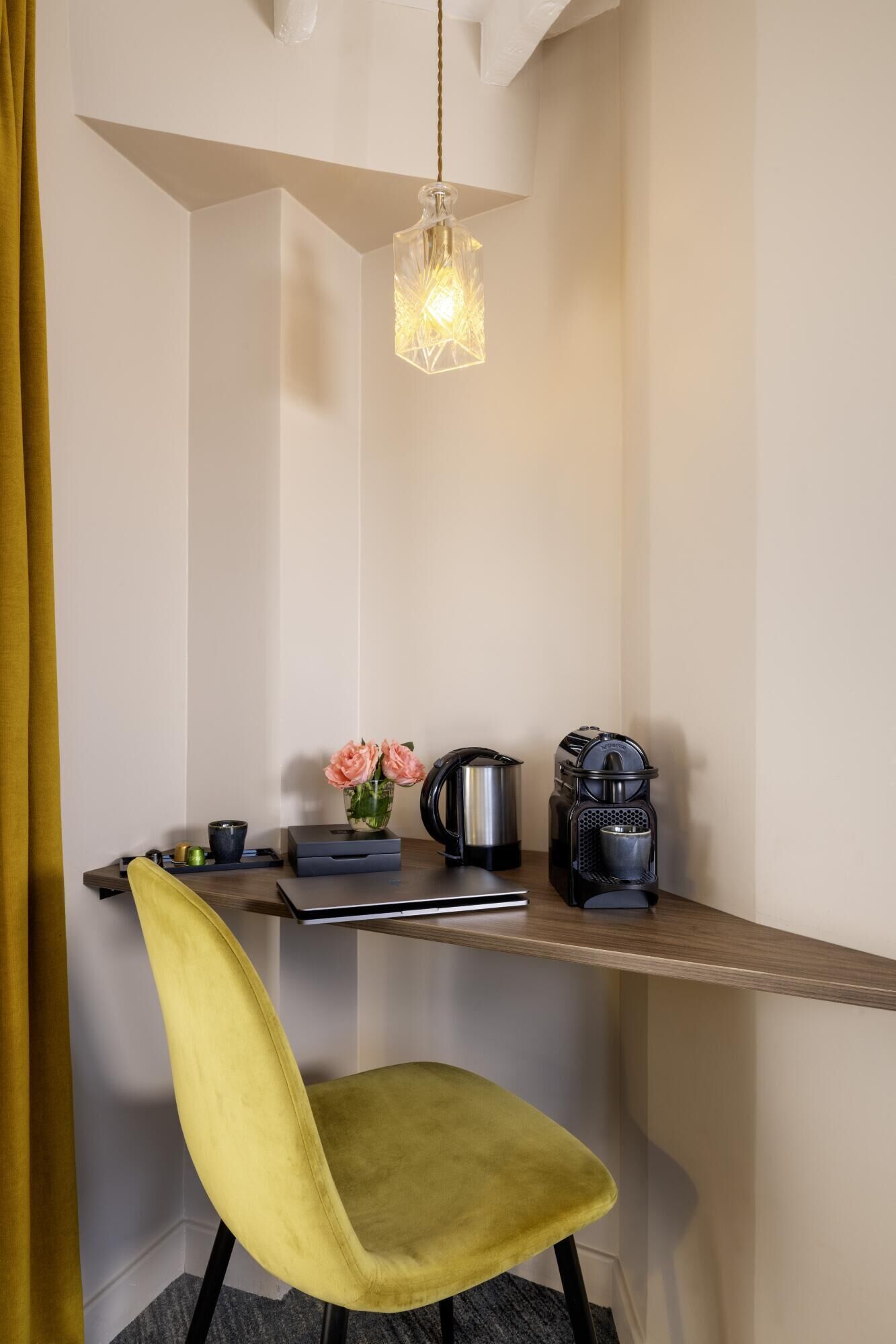 Un espace de travail compact avec une chaise jaune, une machine à café, une bouilloire et une petite table sur laquelle est posé un vase de fleurs.