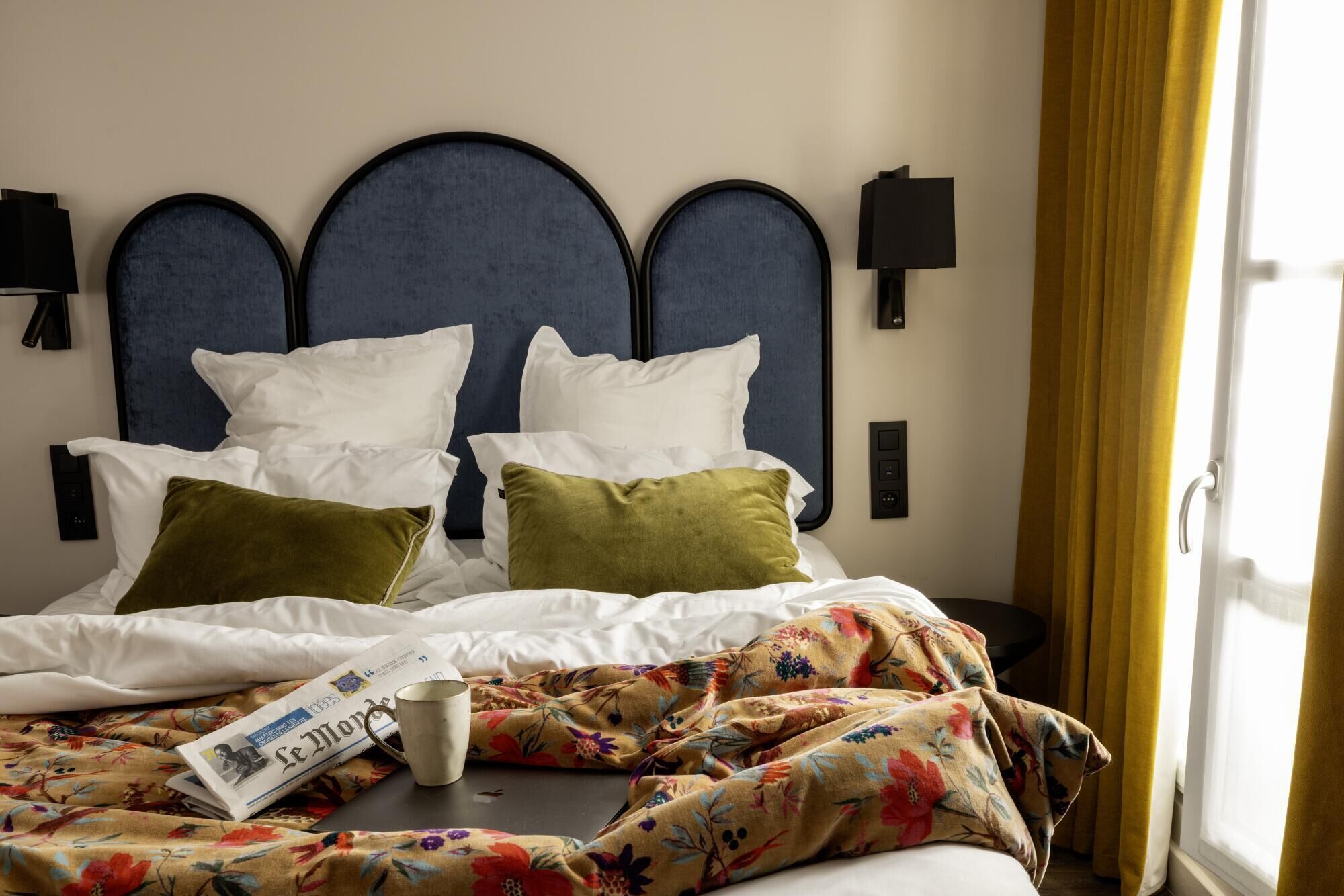 Habitación de hotel con cama, dos almohadas, mesita de noche con una taza y una manta estampada.