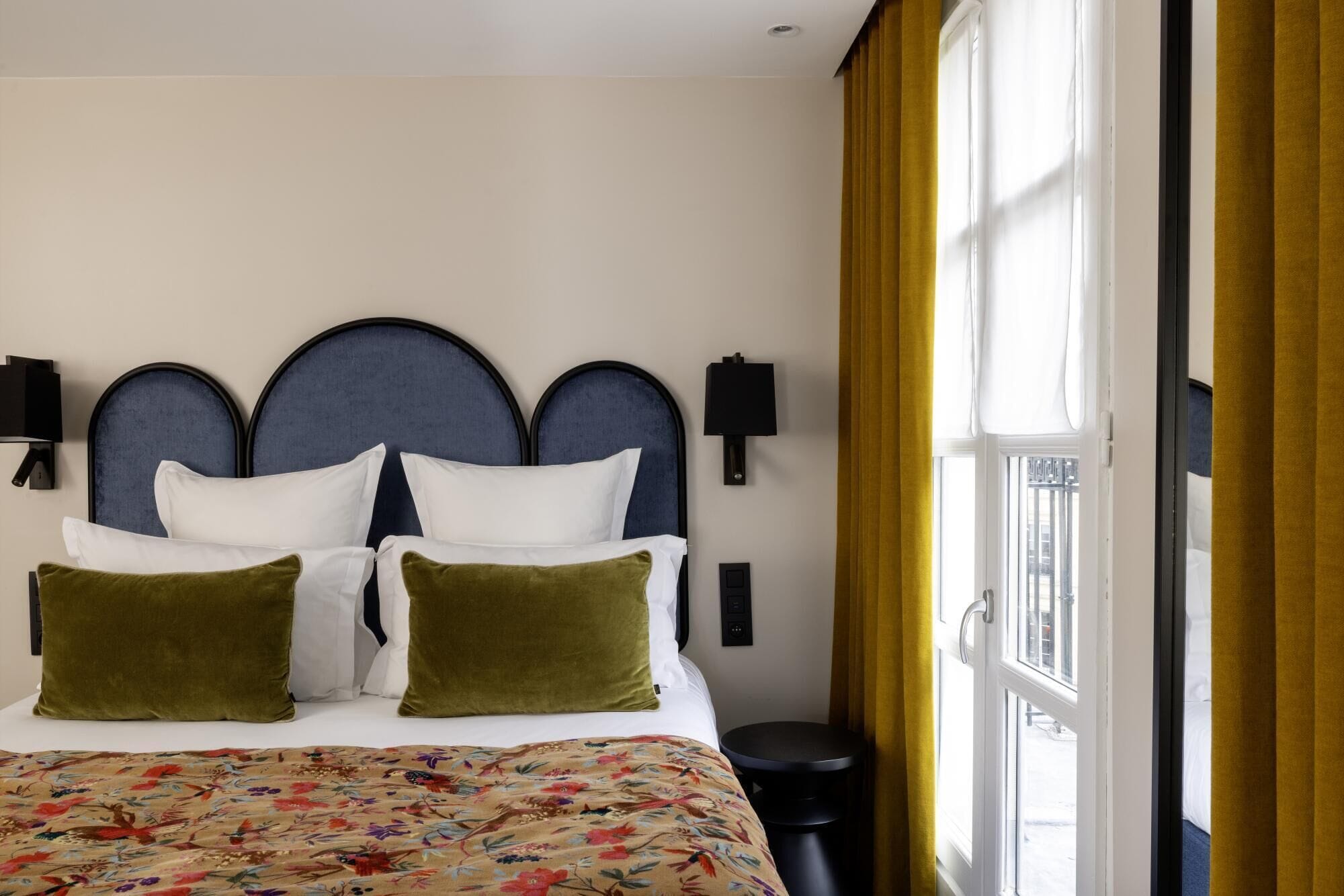 A hotel room with a large bed, two wall-mounted lamps, a window with yellow curtains, and a small round table.
