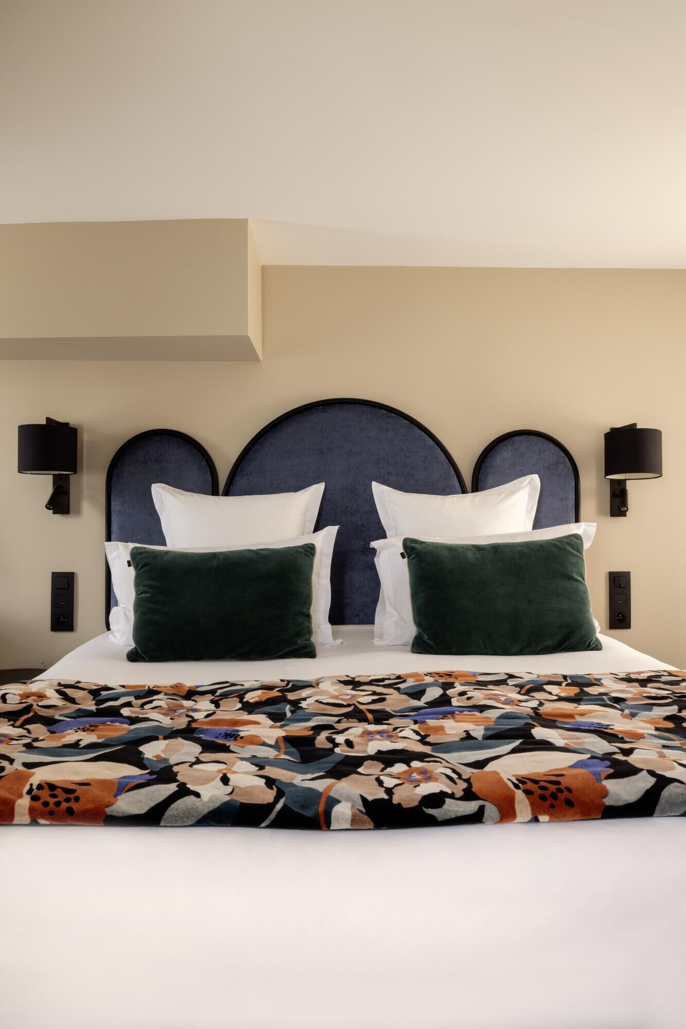 A hotel room with a large bed, two green pillows, a patterned bedspread, and a dark headboard.