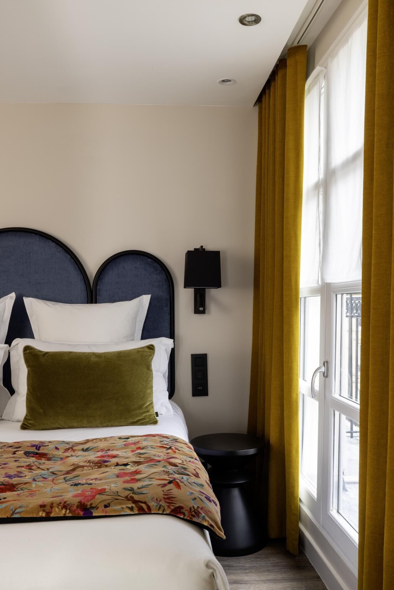 Habitació d'hotel amb un llit, una taula de nit, una làmpada, un safataire i una finestra amb cortines.