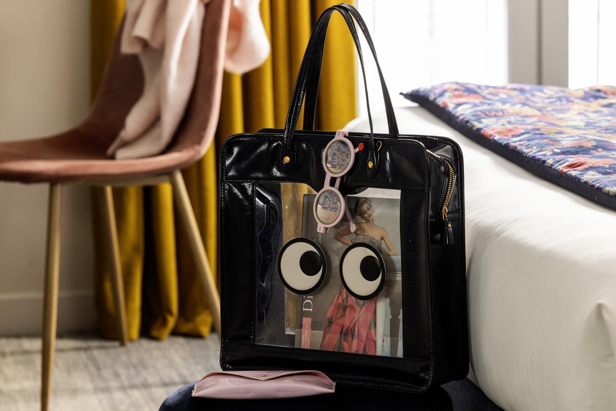 Bolso negro con una ventana transparente y una imagen de una persona en la parte frontal, sobre una cama con edredón estampado con motivos florales.