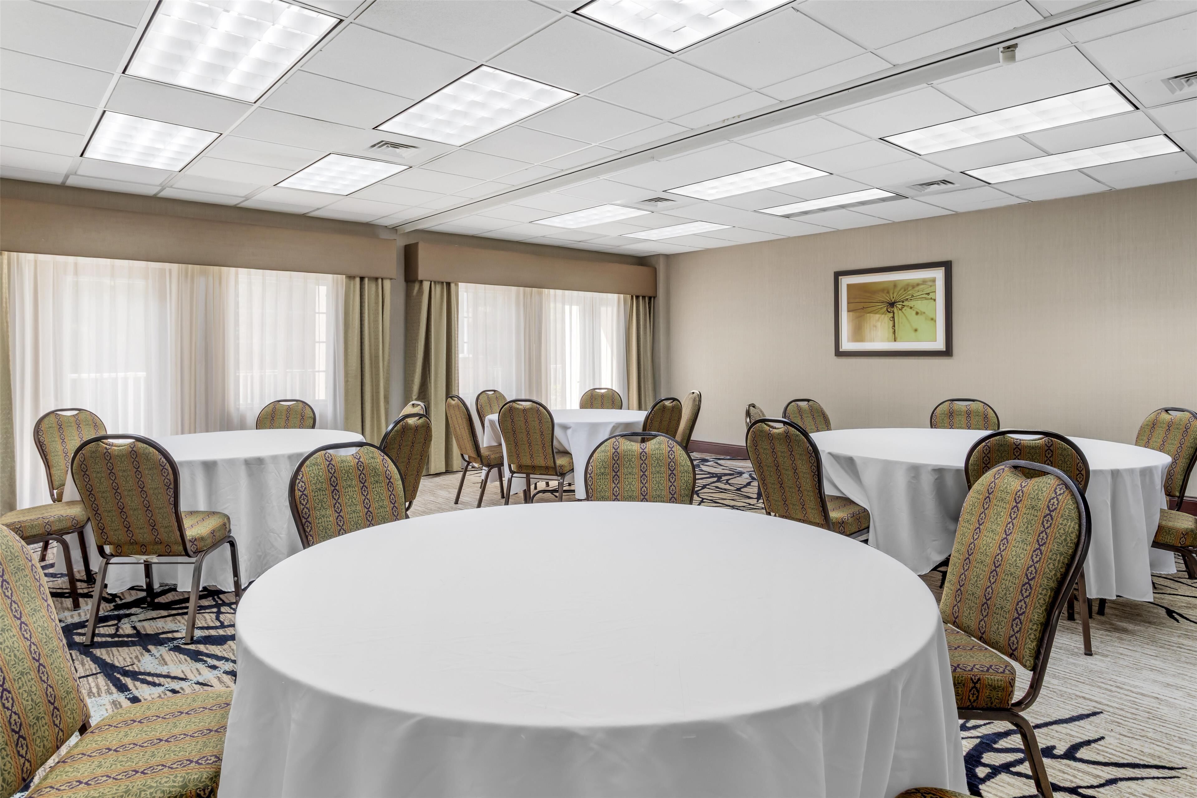 A conference room with round tables and patterned chairs, large windows with curtains, and a framed picture on the wall.