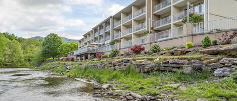 A multi-story hotel with balconies, a river in the foreground, and a mountainous backdrop.
