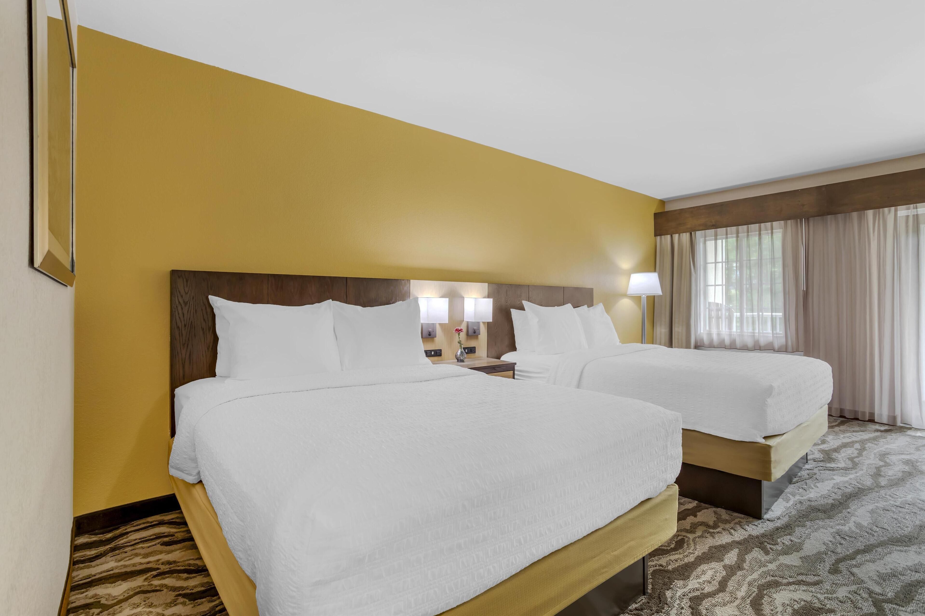 Una habitación de hotel con dos camas, un cabecero de madera, alfombra estampada y una ventana con cortinas.