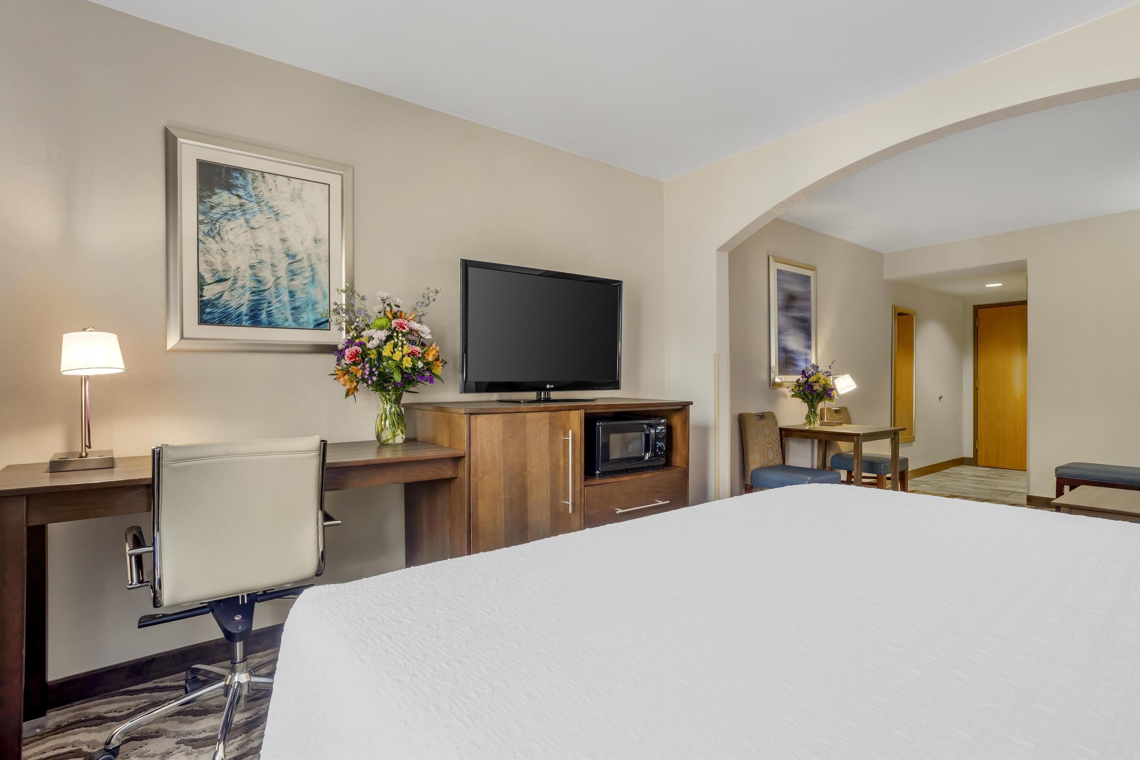 A hotel room with a large bed, a desk with a lamp, a flat-screen TV, a microwave, and a flower vase.