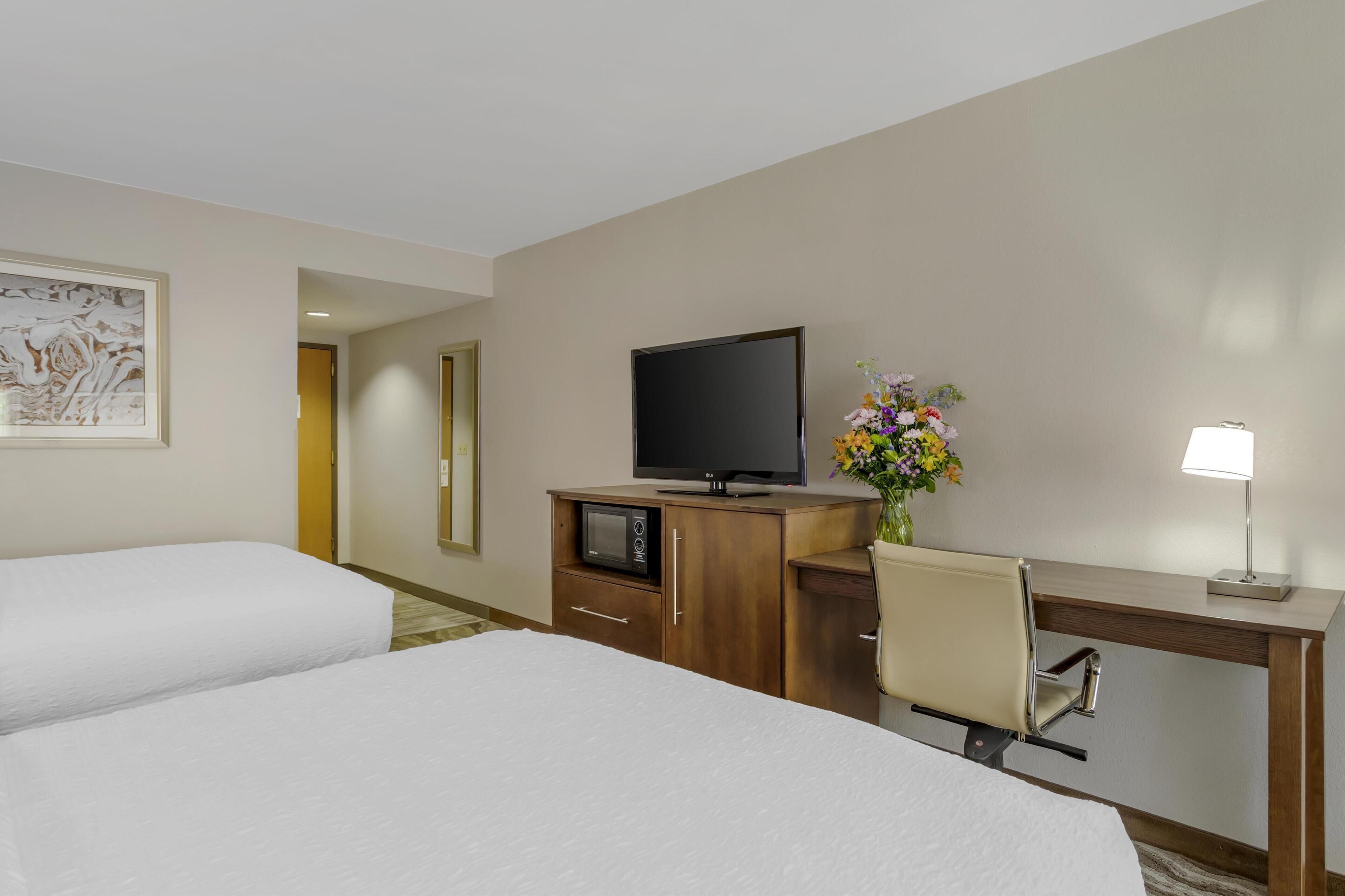 A hotel room with two beds, a desk with a TV, a microwave, a chair, and a lamp.
