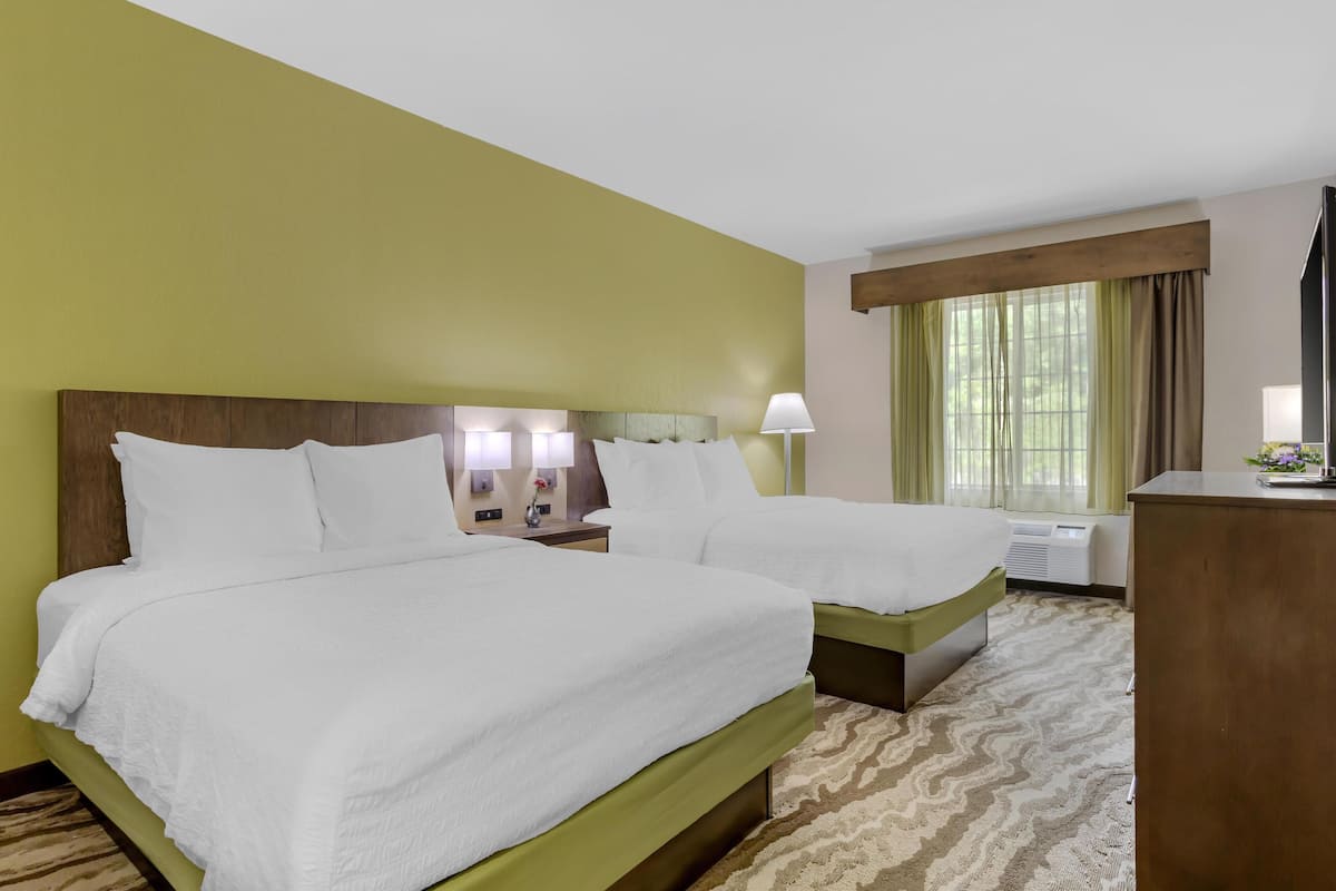 A hotel room with two beds, a wooden headboard, a nightstand with a lamp, a window with curtains, and a desk with a lamp.