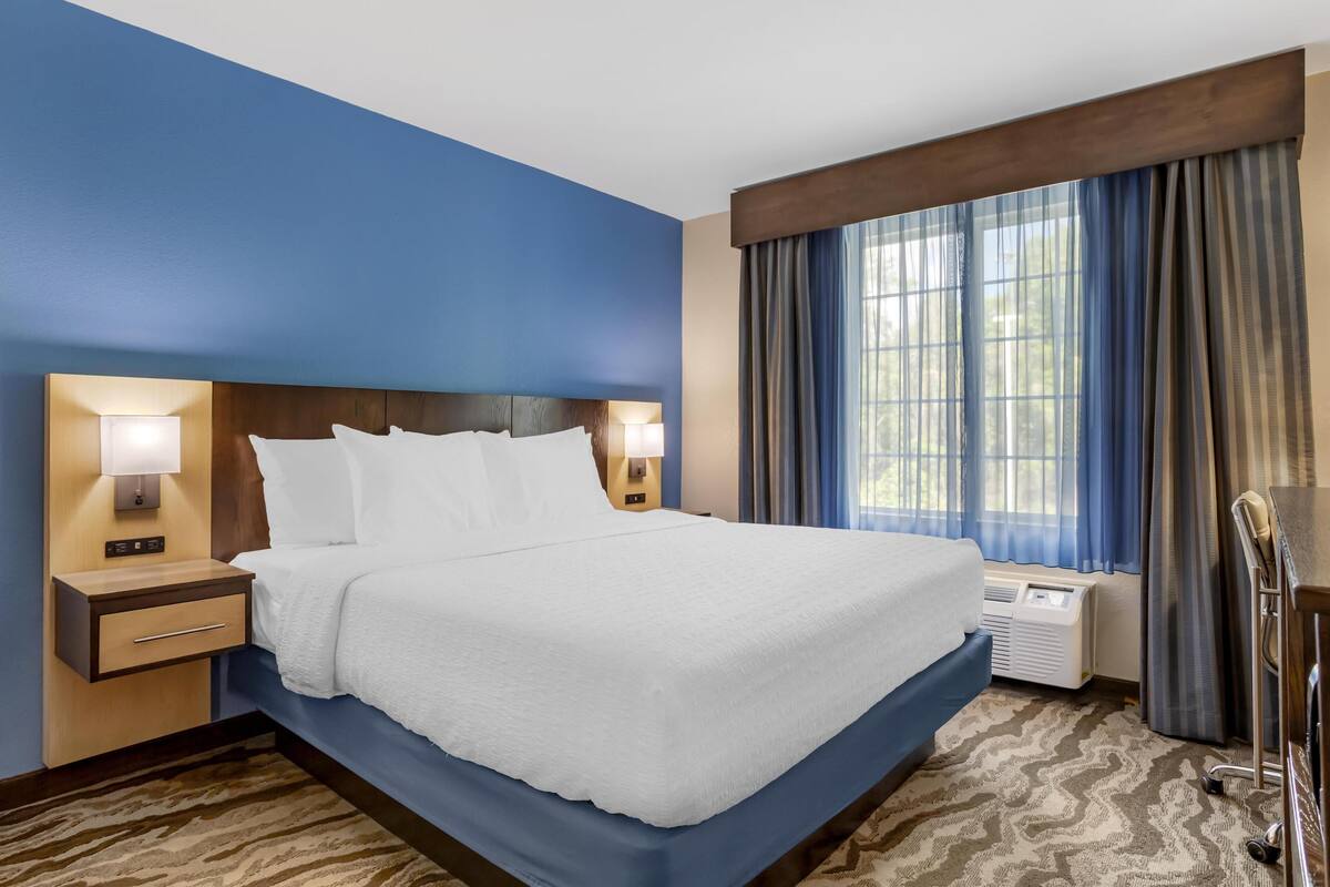 A hotel room with a large bed, two bedside lamps, a window with curtains, and a wall-mounted air conditioning unit.