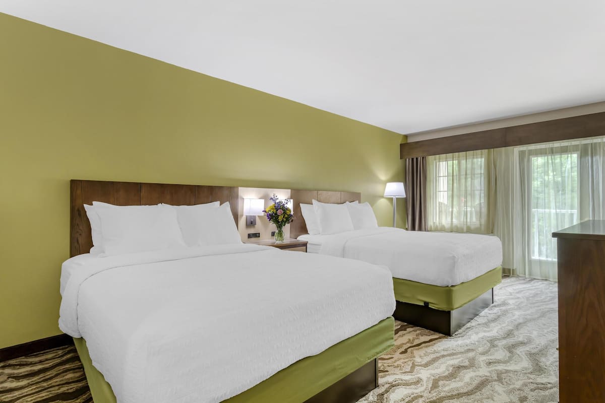 A hotel room with two beds, a nightstand with a lamp, and a view of trees through the window.