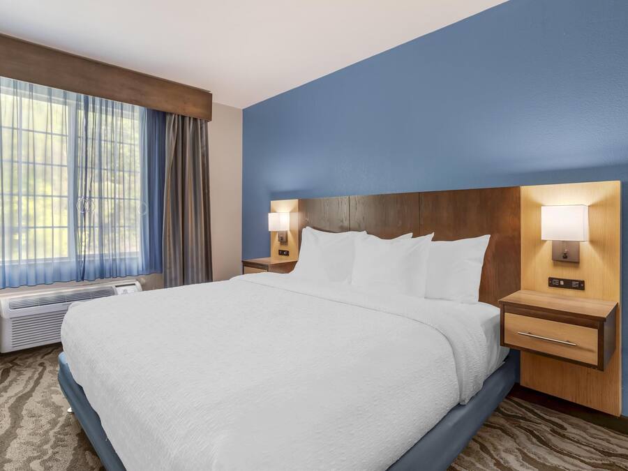 A hotel room with a large bed, two bedside tables with lamps, a window with curtains, and a wall-mounted heater.