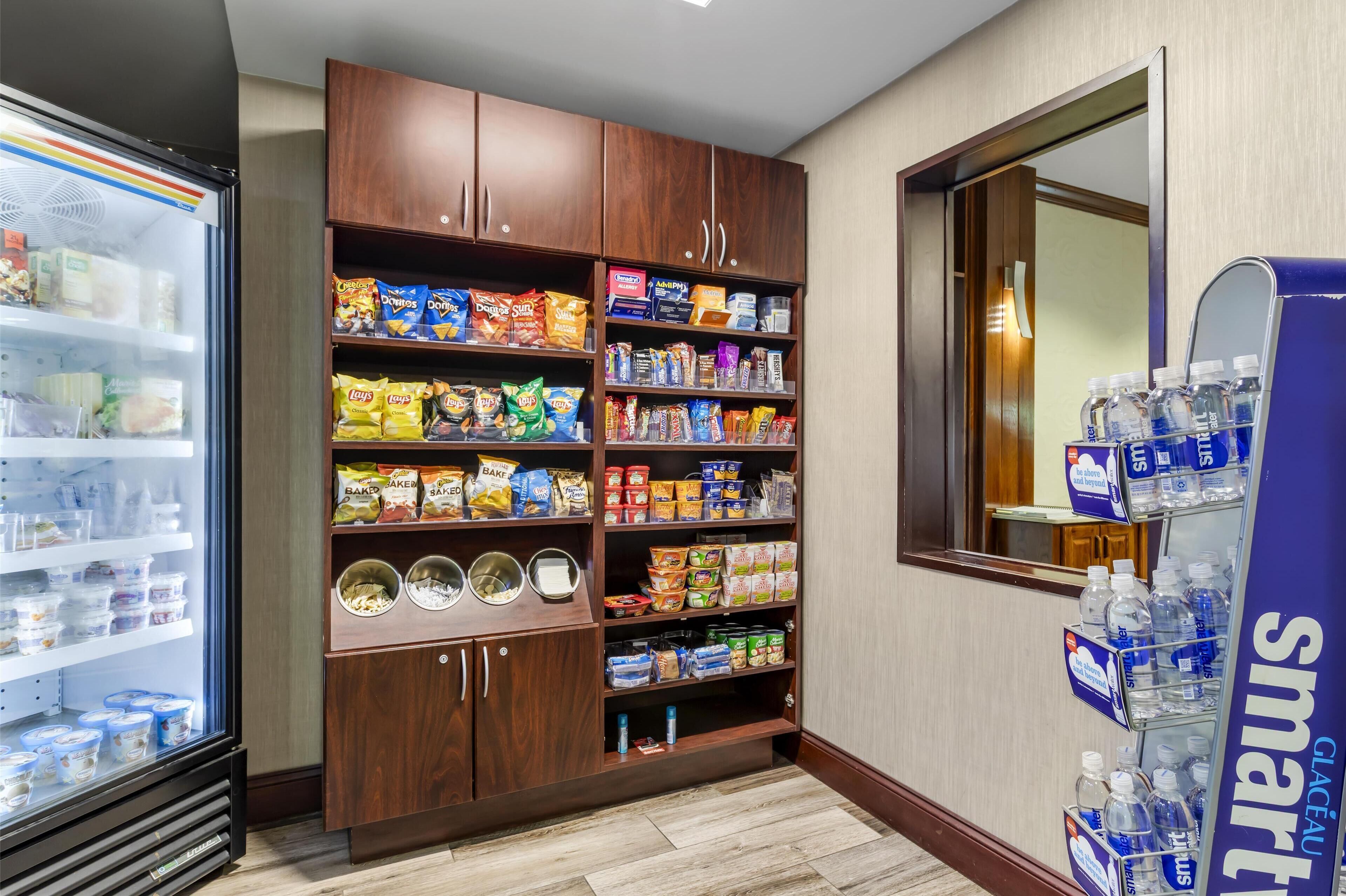 A hotel pantry stocked with snacks and beverages, a refrigerator with dairy products, and a water dispenser.