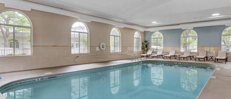 A large indoor swimming pool with clear glass walls, multiple windows, and several lounge chairs.