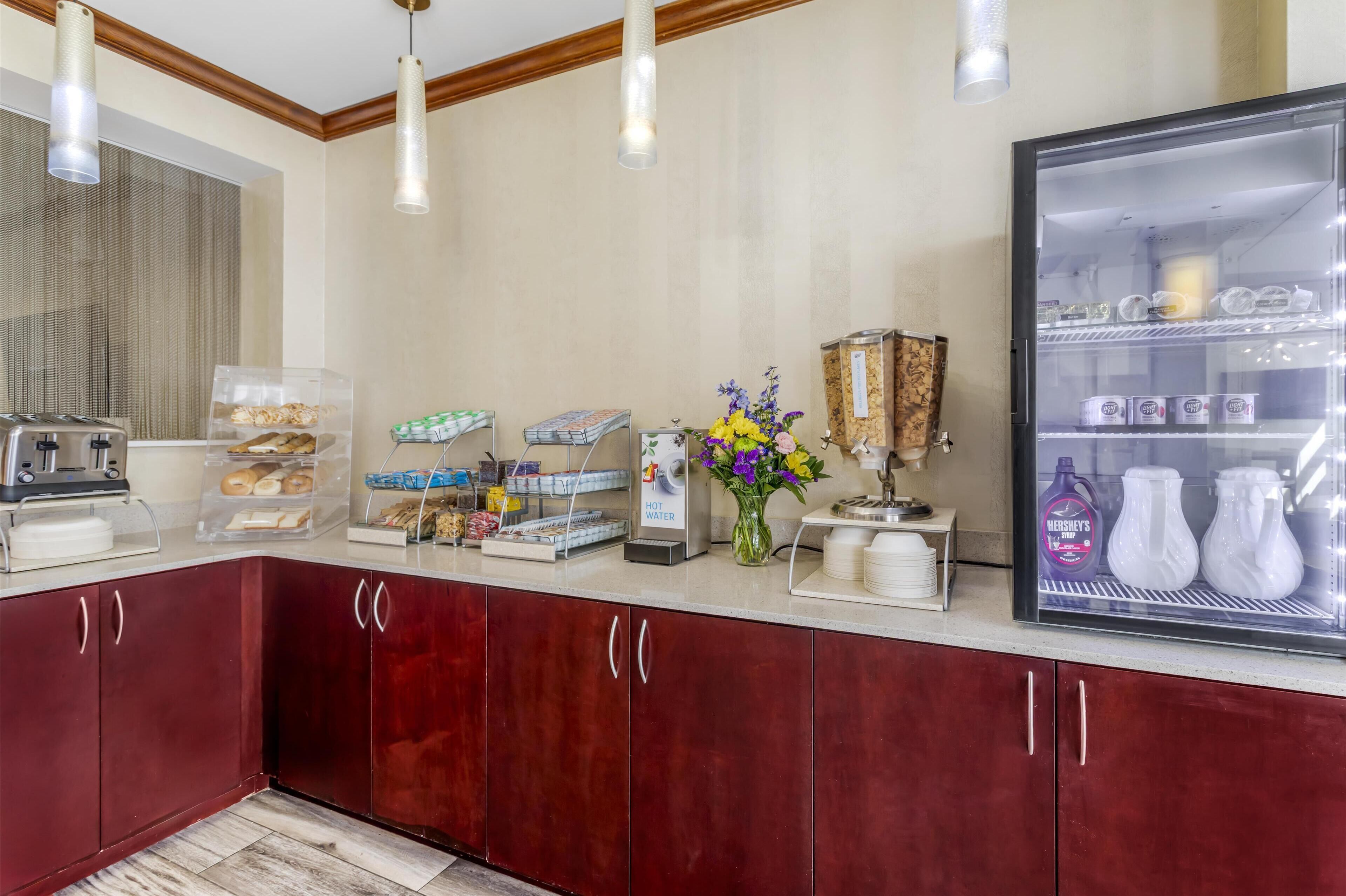 A hotel breakfast area with a refrigerator, coffee machine, and various food items.