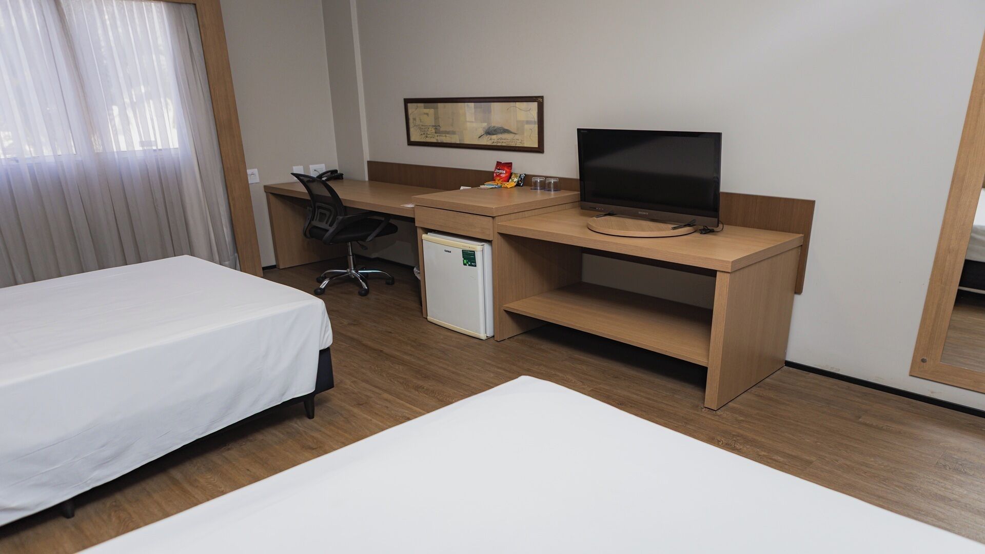 Quarto de hotel com uma cama, uma escrivaninha com televisão, uma cadeira e um pequeno frigorífico.