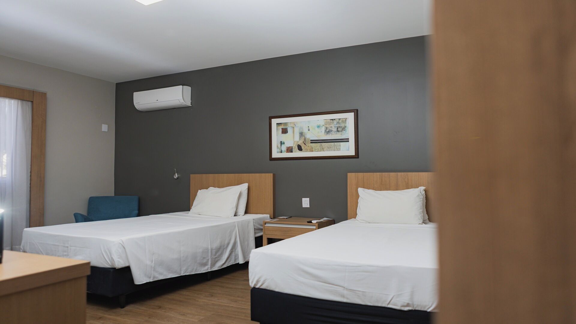 A hotel room with two beds, a desk, a chair, a painting on the wall, and an air conditioning unit.