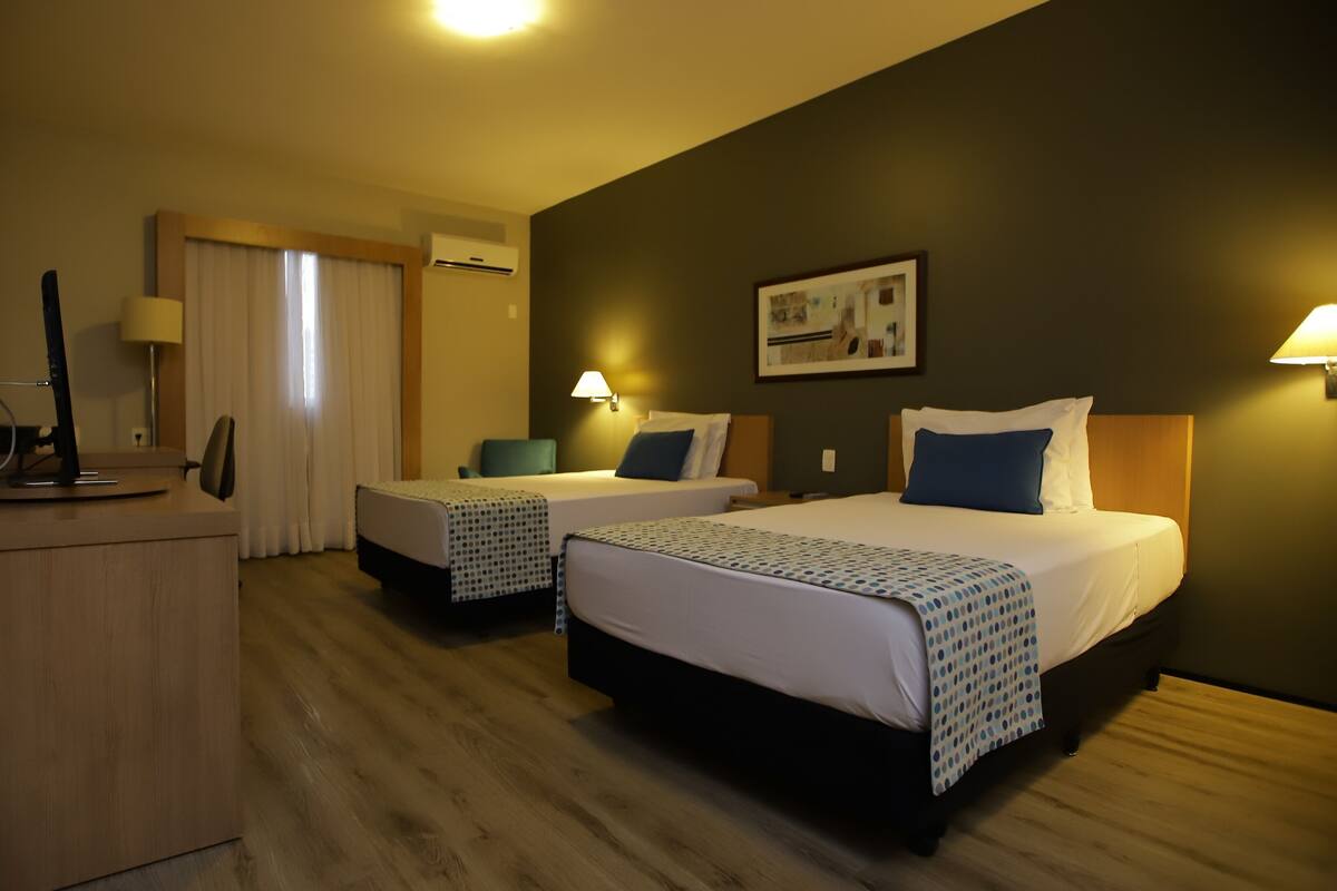 A hotel room with two beds, a desk, a chair, a lamp, and an air conditioning unit.