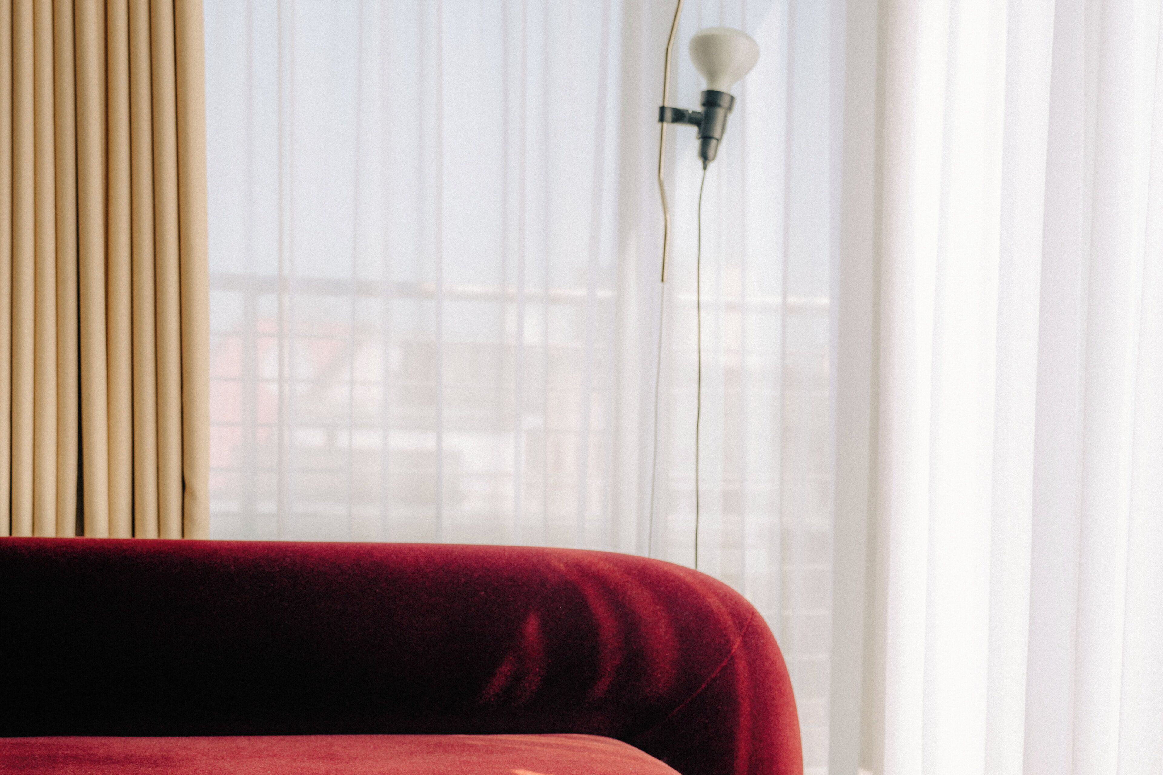 A room with a red upholstered headboard, a window with sheer curtains, and a wall-mounted light fixture.