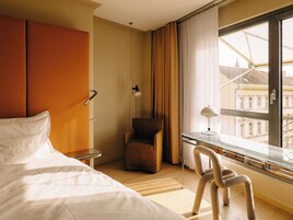 Quarto de hotel com uma cama, uma cadeira, uma escrivaninha e janela com vista para edifícios.