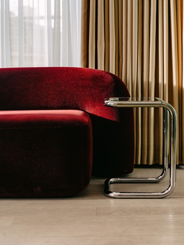 Um sofá de veludo vermelho e uma mesa lateral de metal cromado num quarto com soalho de madeira e cortinas bege.