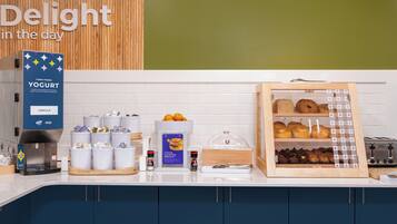 Un buffet de petit-déjeuner comprenant un distributeur de yaourt, une machine à café et une variété de viennoiseries.